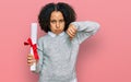 Young little girl with afro hair holding graduate degree diploma with angry face, negative sign showing dislike with thumbs down, Royalty Free Stock Photo