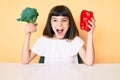 Young little girl with bang holding broccoli and red pepper celebrating crazy and amazed for success with open eyes screaming