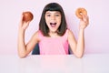 Young little girl with bang holding red apple and donut sitting on the table celebrating crazy and amazed for success with open