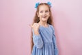 Young little girl standing over pink background smiling with happy face looking and pointing to the side with thumb up Royalty Free Stock Photo