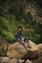 Young lonely tourist cutie girl sitting on stone on shore of river in mountains in wild terrain and satisfying thirst Royalty Free Stock Photo
