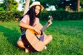 Young long-haired Latina girl, in red and white striped shirt and hat, sitting on a garden lawn playing an acoustic guitar Royalty Free Stock Photo