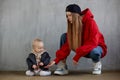 Young long haired mom in red hoodie and black baseball cap plays with her cute baby in beige hoodie and funny motorcycle Royalty Free Stock Photo