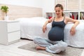 Young long-haired pregnant woman sitting on the mat with her legs crossed and elbows on the bed after yoga exercises, smiling.