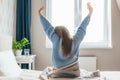 Young long-haired woman outstretching her arms sitting on the bed after good night sleep in light modern bedroom, back Royalty Free Stock Photo