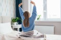 Young long-haired woman outstretching her arms sitting on the bed after good night sleep in light modern bedroom, back Royalty Free Stock Photo