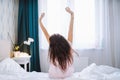 Young long-haired woman outstretching her arms sitting on the bed after good night sleep in light modern bedroom, back Royalty Free Stock Photo