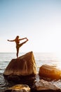 Young long-haired woman in Utthita Padangushthasana from yoga standing on a big stone in water. Sunset, relaxation