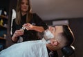 Young longhaired female Barber gently applies shaving cream foam on bearded man's face bristle while he lying in barbershop Royalty Free Stock Photo