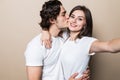 Young lovely couple posing together with klosed eyes while making selfie and man kissing woman over beige background Royalty Free Stock Photo