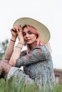 Young lovely woman with pink hair in vintage straw hat in stylish summer blue dress sits on fresh green grass outdoors on nature. Royalty Free Stock Photo