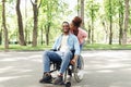 Young loving black woman kissing her happy boyfriend in wheelchair on walk at city park, full length Royalty Free Stock Photo