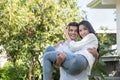 Young loving couple have fun in park. Caucasian boyfriend carry on his asian girlfriend in arms in front yard of house