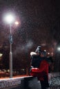 Young loving couple stand near a side of an ice skating rink under a lamp at night, there is a heavy snowfall Royalty Free Stock Photo