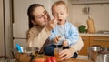 Young loving mother feeding her little baby boy playing with cookware on kitchen. Concept of little chef, children Royalty Free Stock Photo