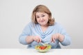 Young lush fat woman in casual blue clothes on a white background at the table and eats a vegetable salad with tomatoes Royalty Free Stock Photo