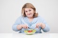 Young lush fat woman in casual blue clothes on a white background at the table and eats a vegetable salad with tomatoes Royalty Free Stock Photo