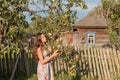 A young maiden woman stands near the garden fence in front of an old village house and closes her eyes in the rays of the morning