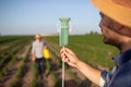 Young male agronomist holding up rain gauge with farmer using hand sprayer in background