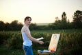 Young male artist, standing by easel, drawing portrait on canvas on green field in summer during sunset. Painting workshop in