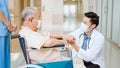 Young male Asian doctor crouch down holding hand and talk to the senior adult patient on wheelchair in hospital hallway Royalty Free Stock Photo