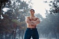 Young bodybuilder with bare torso stands with arms crossed in winter misty forest. Royalty Free Stock Photo