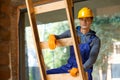 Young male builder in overalls and hard hat looking at camera, climbing up the ladder while working at cottage