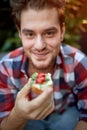 Young male caucasian holding sandwich that is about to eat, looking at camera, smiling