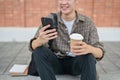 A young male college student sits on the street using his smartphone and holding a coffee cup