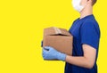 Young male courier in a medical mask and rubber gloves holding a box on a yellow background. Social concept. Helping the poor.