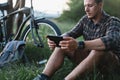 Young Male Cyclist Sits Resting On Summer Meadow Near Bicycle, Holding At Tablet Recreation Resting Travel Destination Concept