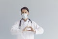 Young male doctor in medical mask showing heart gesture on grey background. Cardiovascular health, charity or donation