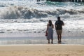 Young male and female couple are seen looking out at the sea on the beach at the edge of a rough ocean Royalty Free Stock Photo