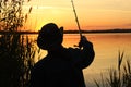 A young male fisherman in a hat throws a fishing rod into the river at sunset. Silhouette of a man in a hat with a fishing rod in Royalty Free Stock Photo