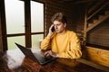 Young male freelancer works in a wooden country house on a laptop at a table and calls on a smartphone, looking at the screen with Royalty Free Stock Photo