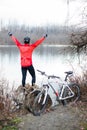 Young male mountain biker standing by the river or lake with arms raised in the air, celebrating success Royalty Free Stock Photo