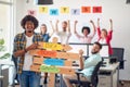 A young male office worker is holding a sign with company slogans and posing for a photo in the office with his colleagues.