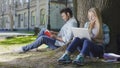 Young male sitting under tree with book near female with laptop, student life