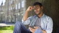 Young male sitting under tree, reading information on cellphone, upsetting news