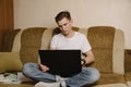 A young male student sits at home on the couch and does homework on a laptop and writes in a notebook.Distance learning Royalty Free Stock Photo