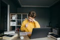 Young male student studying at home with books and laptop on bedroom background, looking at notebook with serous face and biting