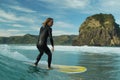 A young male surfer in a black wetsuit rides a longboard surfboard on a wave in front of Lion Rock, Piha Beach, New Zealand