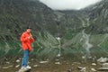 Young male tourist in red jacket stands on a rock in the mountains and uses a smartphone against a background of a mountain lake. Royalty Free Stock Photo