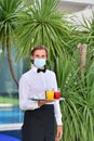 Young male waiter with a surgical mask holding a bar tray with assorted drinks outdoors on an out of focus background. Serving and