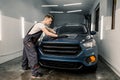 Young male worker washes a car hood, wiping water with a soft microfiber cloth, cleaning the surface to shine in a Royalty Free Stock Photo