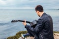 Young man with acoustic guitar playing on beach surrounded with rocks on rainy day Royalty Free Stock Photo