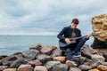 Young man with acoustic guitar playing on beach surrounded with rocks on rainy day Royalty Free Stock Photo