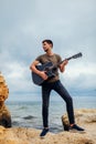 Young man with acoustic guitar playing on beach surrounded with rocks on rainy day Royalty Free Stock Photo