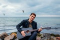 Young man with acoustic guitar playing and singing on beach surrounded with rocks on rainy day Royalty Free Stock Photo