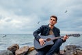 Young man with acoustic guitar playing and singing on beach surrounded with rocks on rainy day Royalty Free Stock Photo